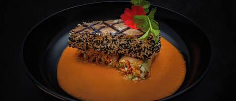 A dish with a fish fillet topped with sesame seeds and a red flower garnish, served on a black plate with a smooth, orange sauce.