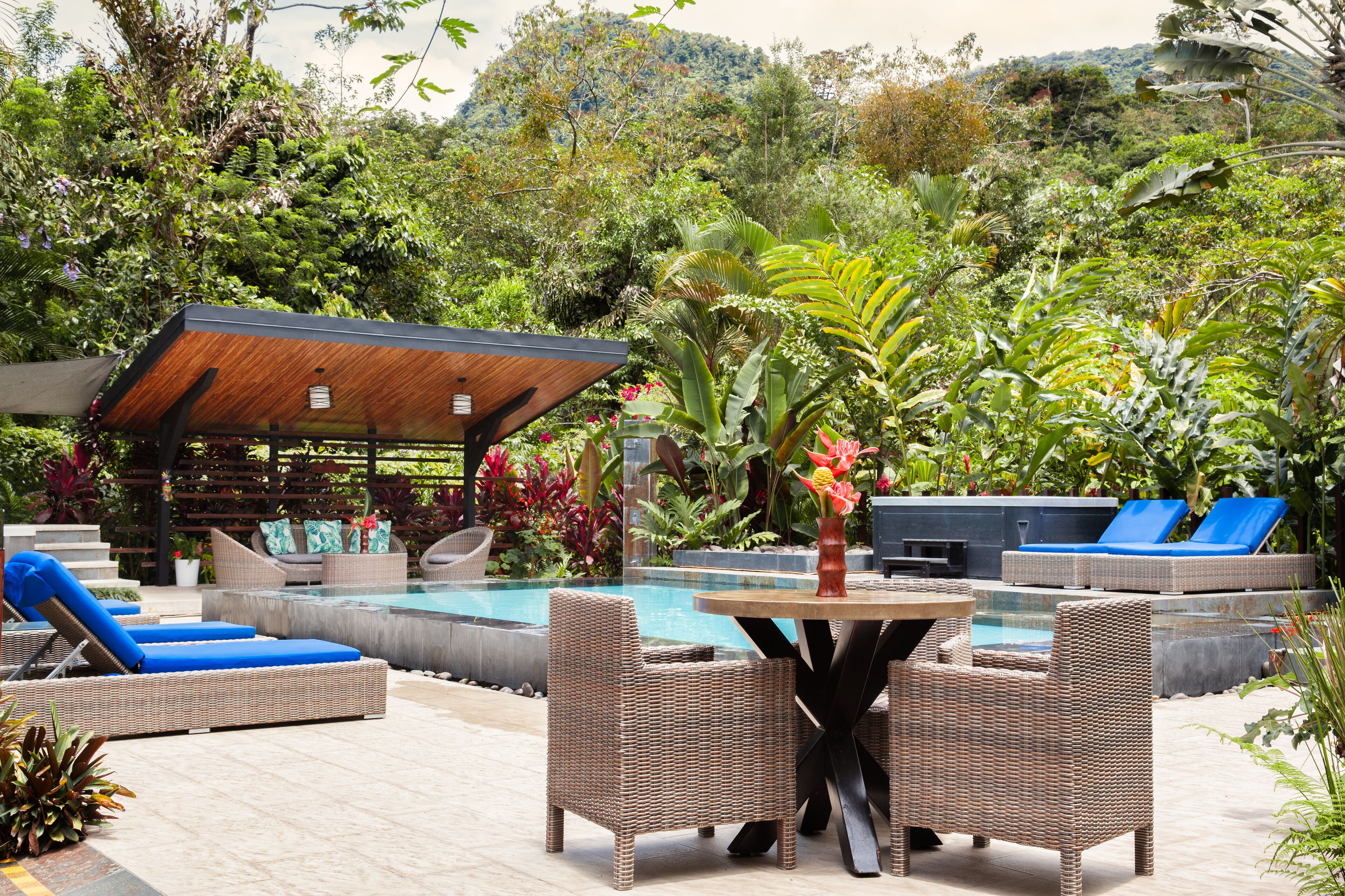 Un espace aménagé au bord de la piscine, avec des meubles en osier, une pergola en bois et une végétation luxuriante.