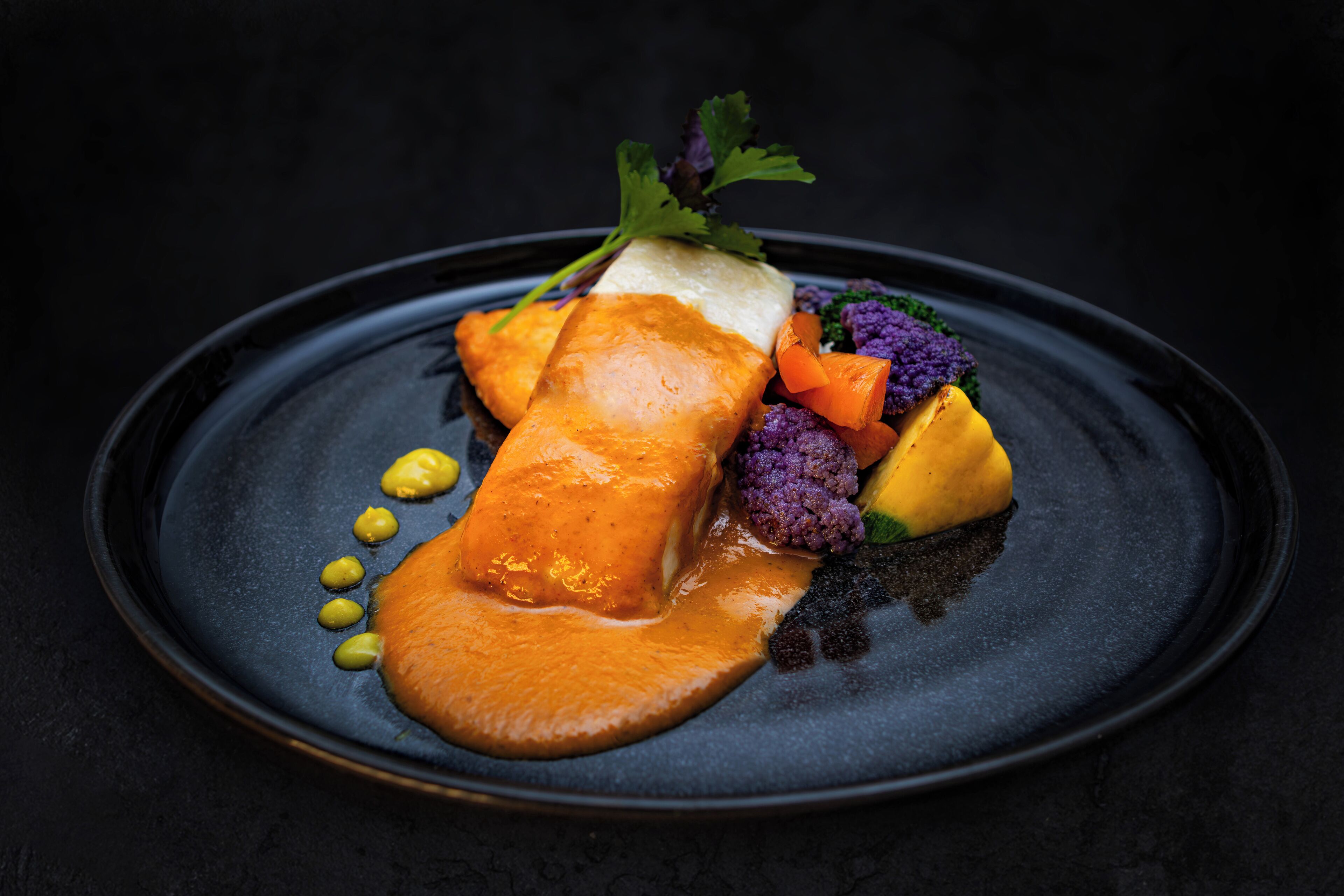 A dark plate with a salmon fillet, vegetables, and a garnish.