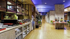 Un moderno vestíbulo de hotel con un mostrador de recepción alargado, una zona de buffet con diversas opciones de comida y un área de descanso con mesas y sillas.