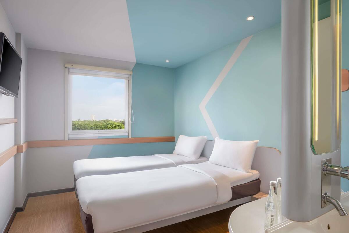 Ein Hotelzimmer mit zwei Betten, einem Fenster mit Blick, einem Badezimmer mit Waschbecken und Spiegel sowie einem flachbildfernseher an der Wand.