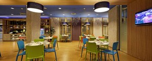 Ein Restaurant mit runden Tischen, bunten Stühlen, einem Fernseher an der Wand und einer Theke mit verschiedenen Artikeln.