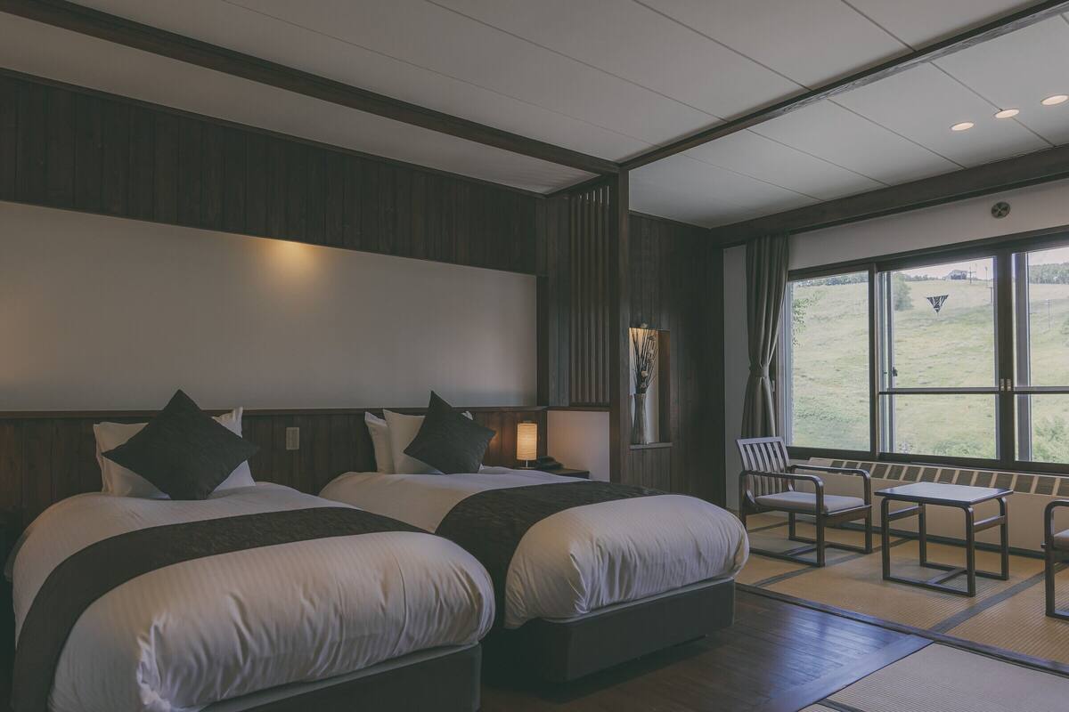 A hotel room with two beds, a small table, and chairs. There is a window with a view of the outdoors.