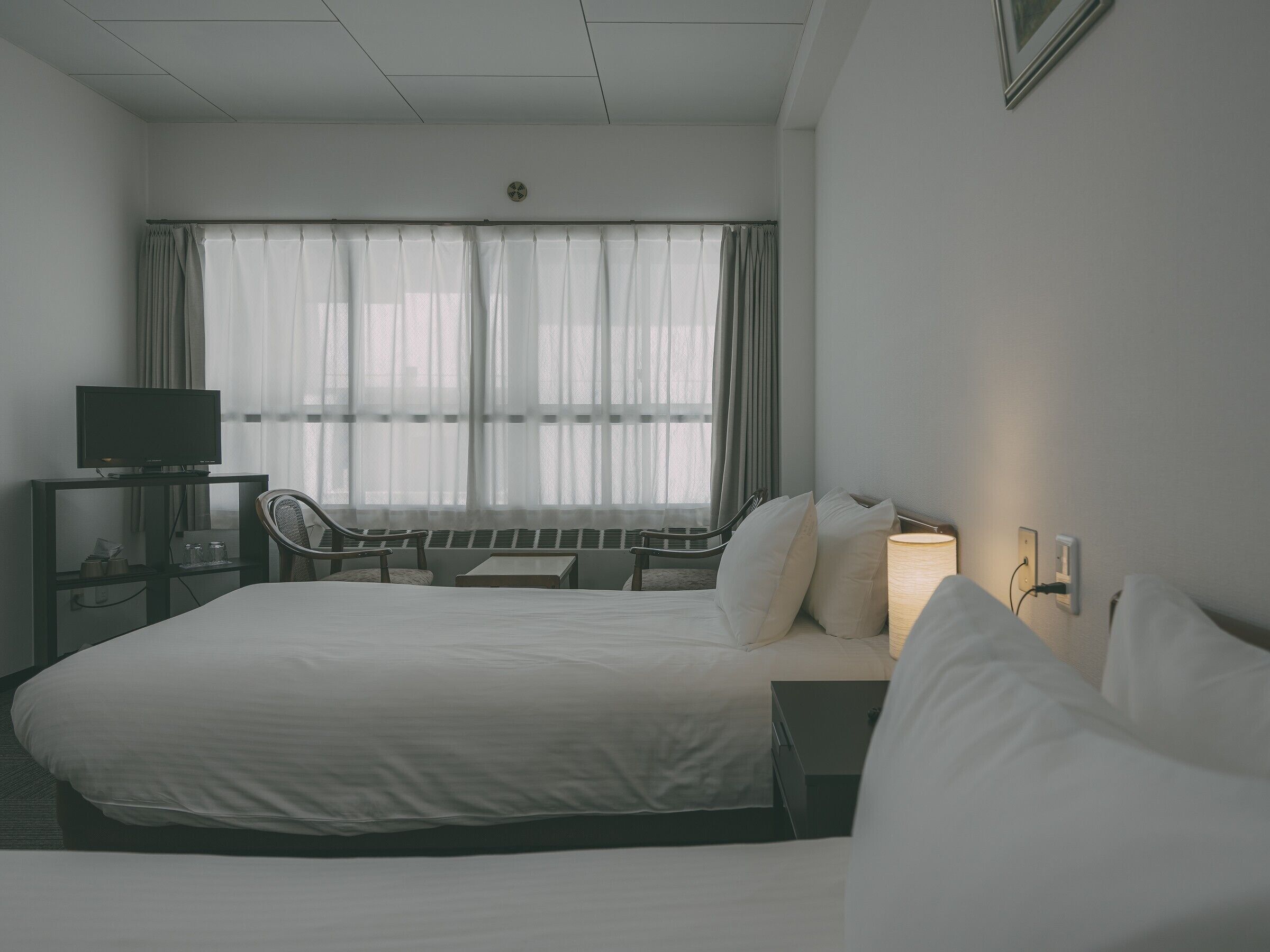 A hotel room with a large bed, a desk with a television, a chair, and a window with sheer curtains.