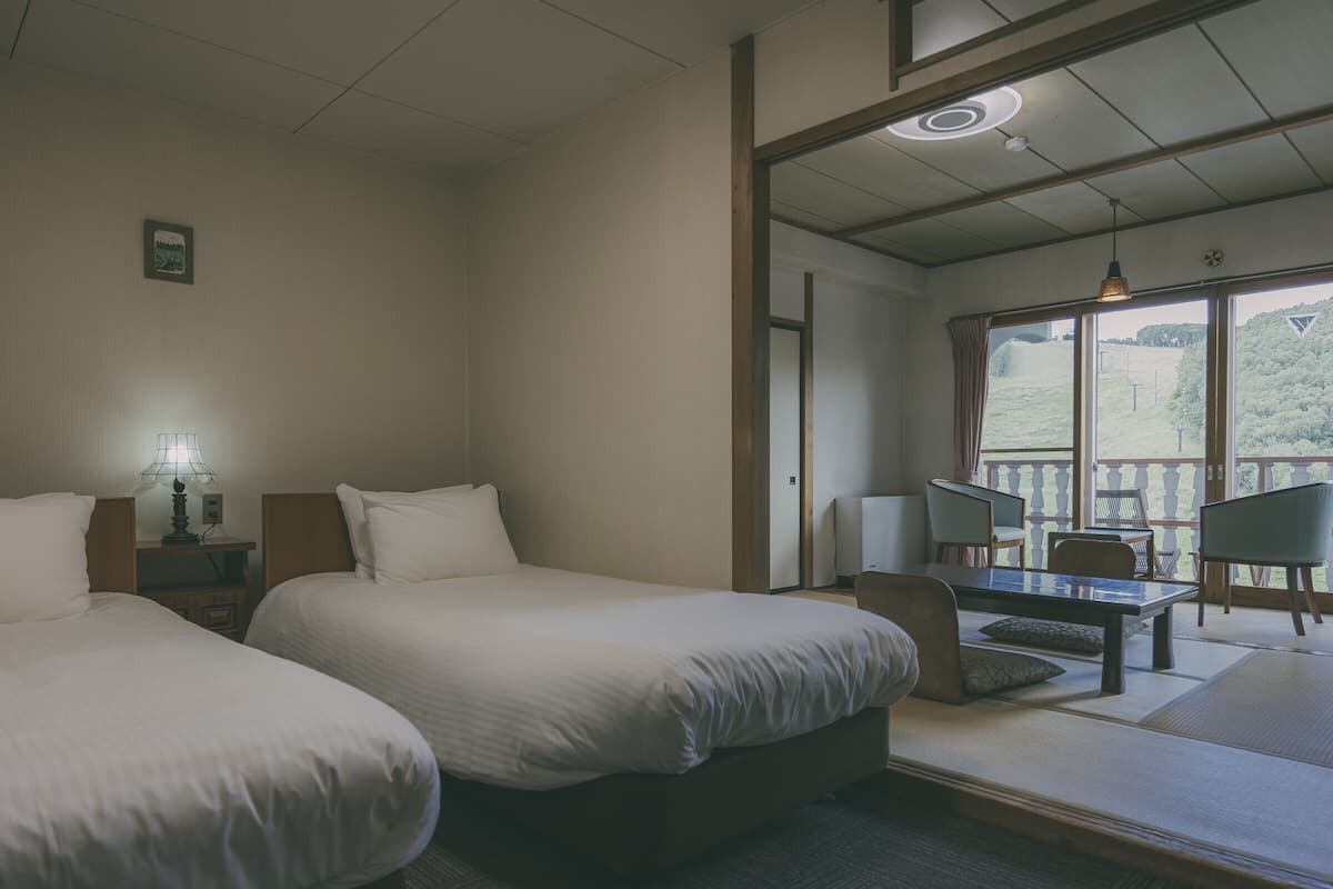 A hotel room with two beds, a desk, chairs, and a view of the outdoors.