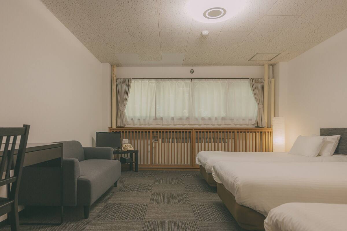 A hotel room with a bed, a desk with a chair, a radiator, and a window with curtains.