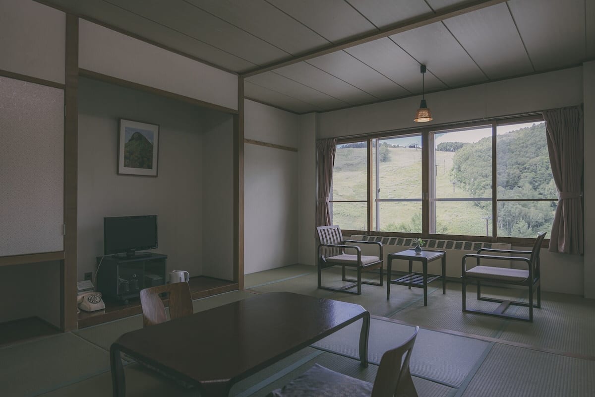 A room with tatami mats, a low table, chairs, a television, and a large window with a view of greenery.