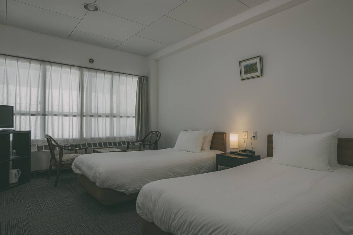 A hotel room with two beds, a desk, a chair, a TV, and a window with curtains.