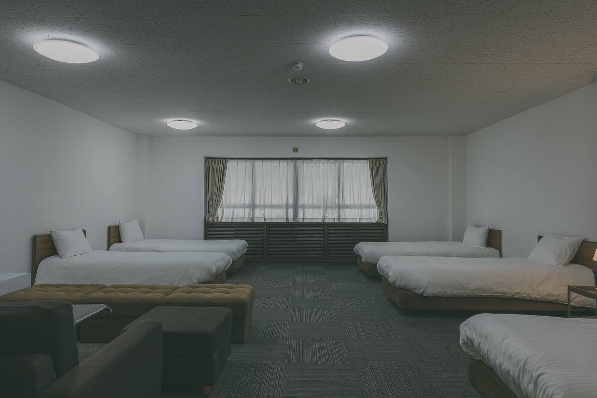 A hotel room with two single beds, a sofa, a window with curtains, and a ceiling with recessed lighting.