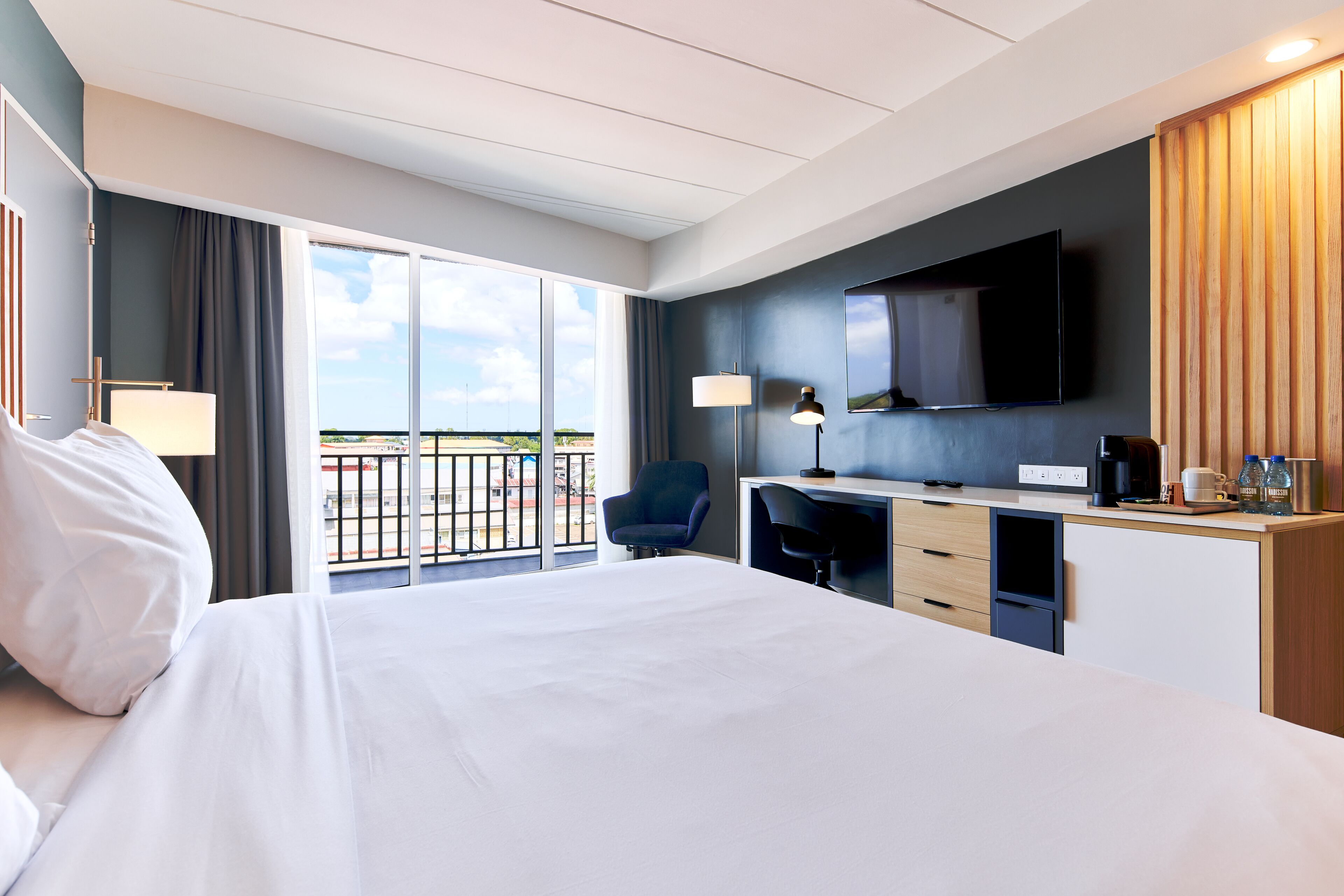 A hotel room with a bed, desk, chair, TV, and a balcony with a view.