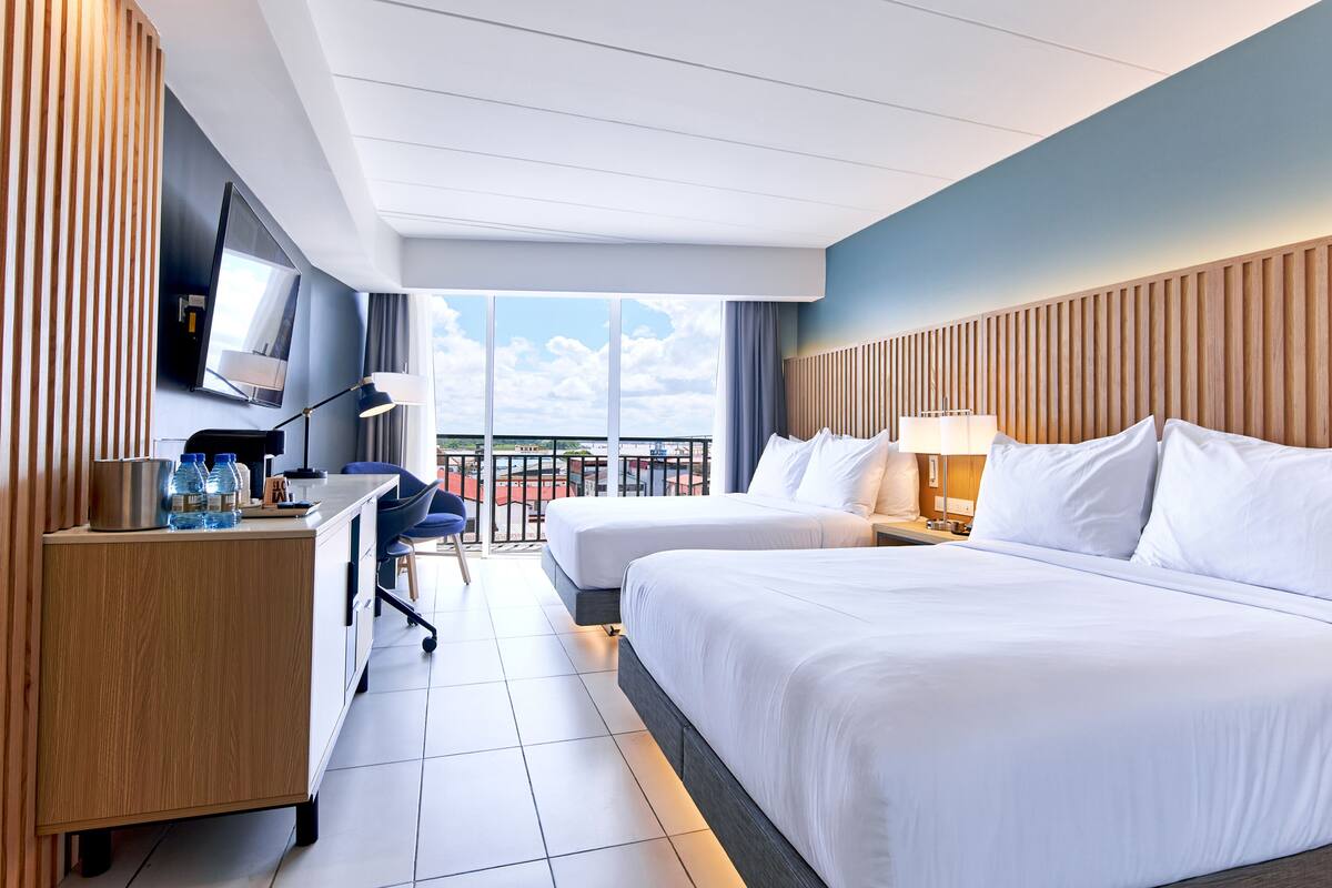 A hotel room with two beds, a desk with a chair, a TV, and a balcony with a view.