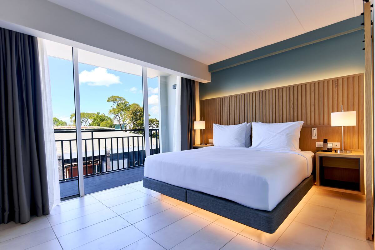 A modern hotel room with a large bed, a balcony with a view, and a wooden headboard.
