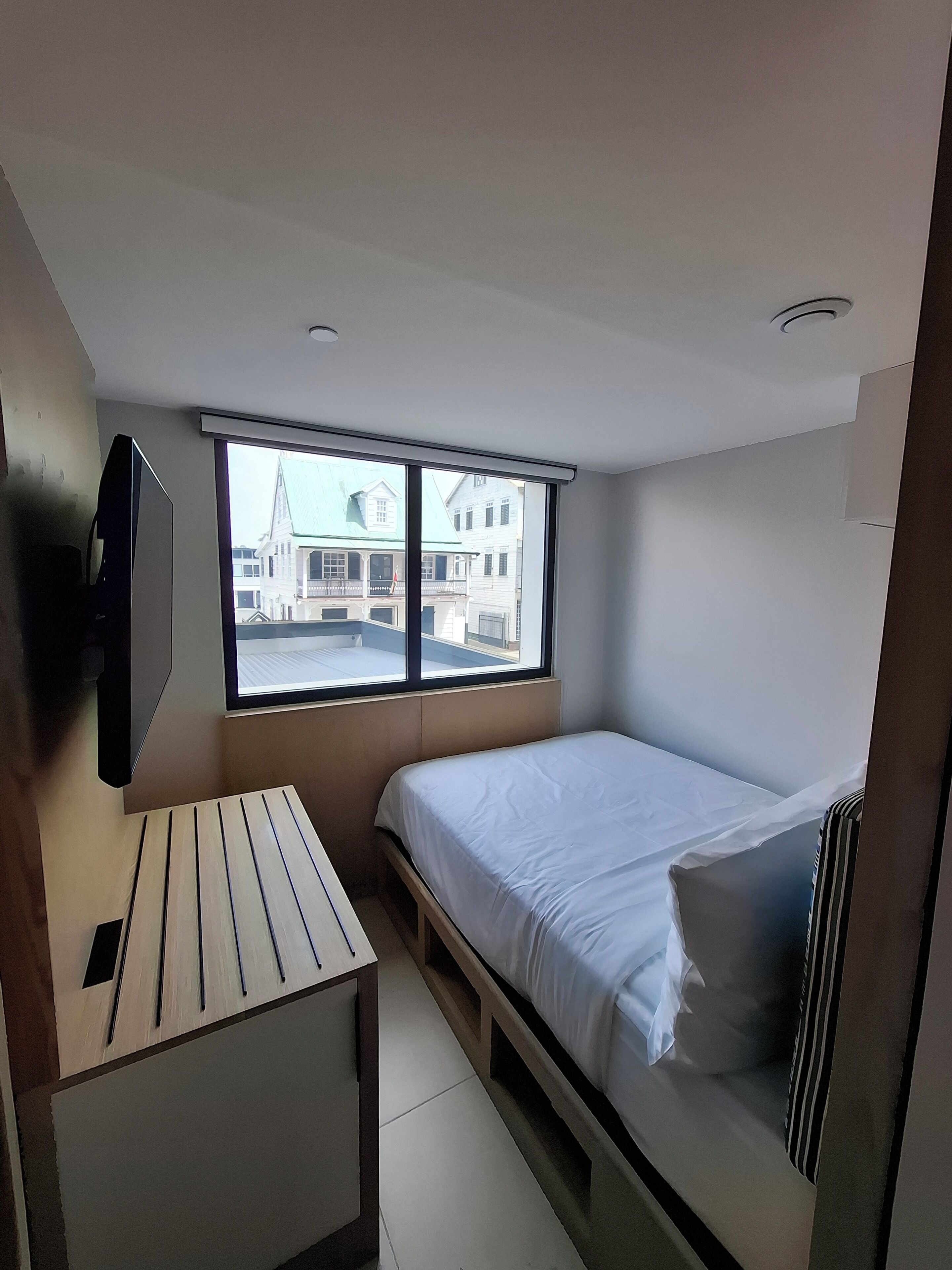 Ein kleiner, moderner Raum mit einem Bett, einem an der Wand montierten Fernseher, einem Fenster mit Blick auf Gebäude und einem kleinen Tisch.