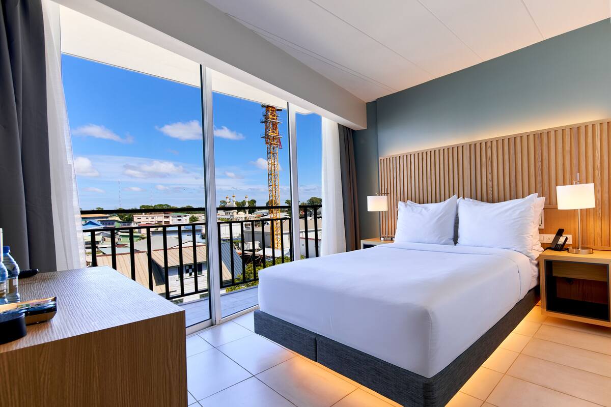 A hotel room with a large bed, a desk, a nightstand, and a view of a cityscape through large windows.