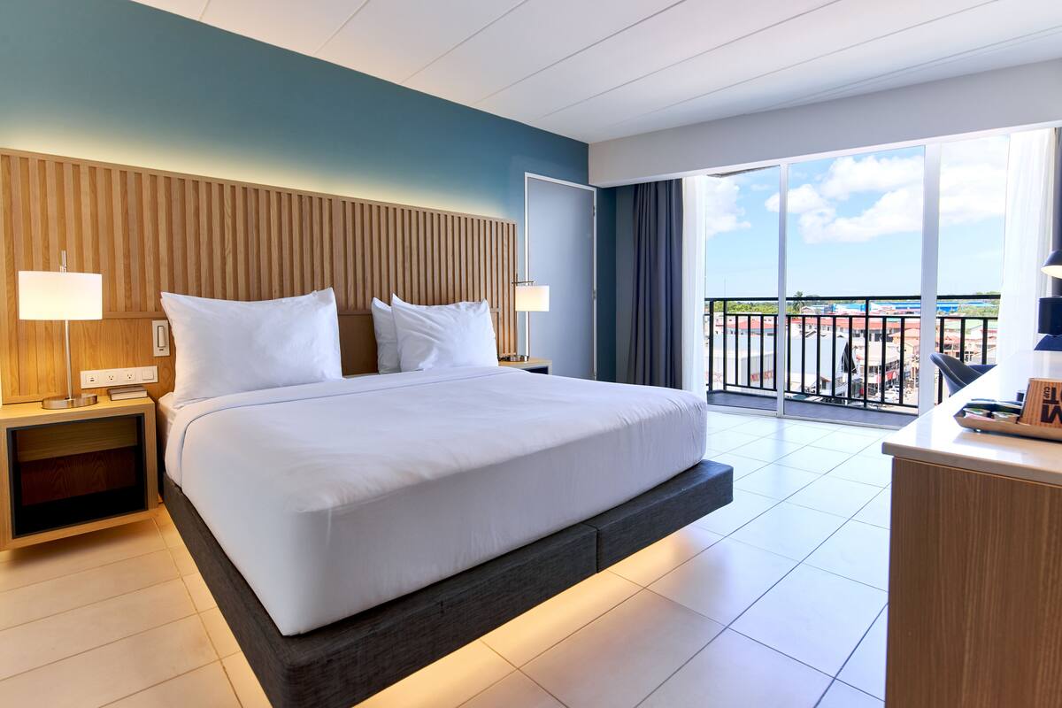A modern hotel room with a large bed, wooden headboard, bedside tables with lamps, a desk, and a balcony with a view.
