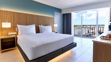 A hotel room with a large bed, a desk, a nightstand, a lamp, and a balcony with a view.