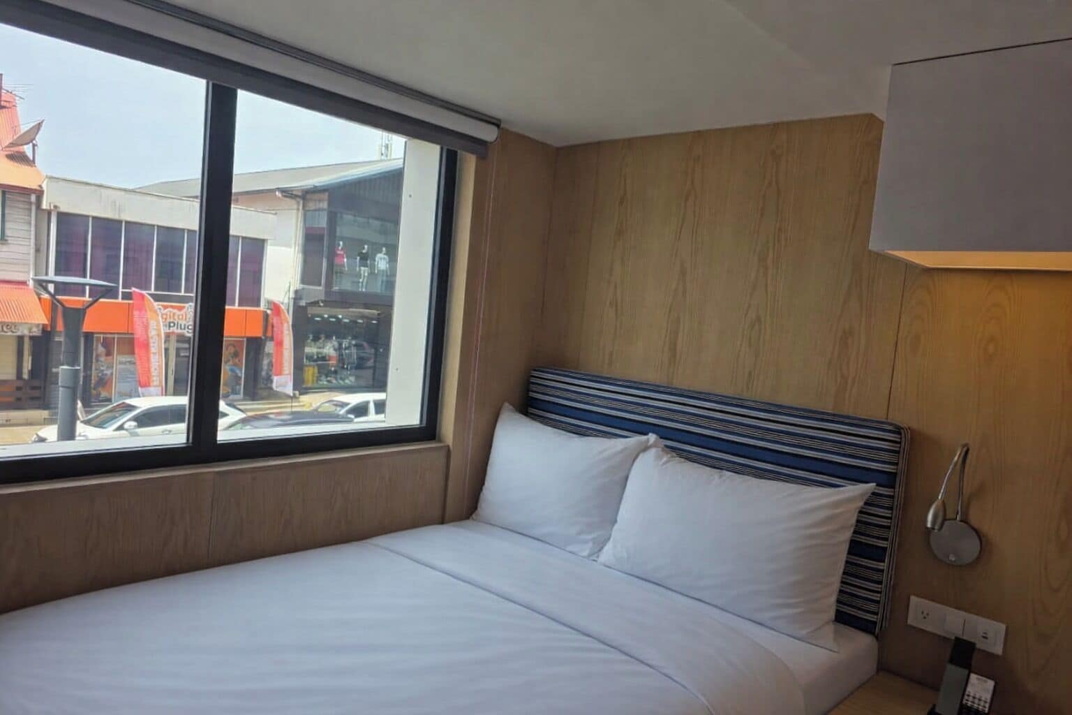 A hotel room with a large window, a bed with white linens, a striped headboard, and a wooden wall panel.