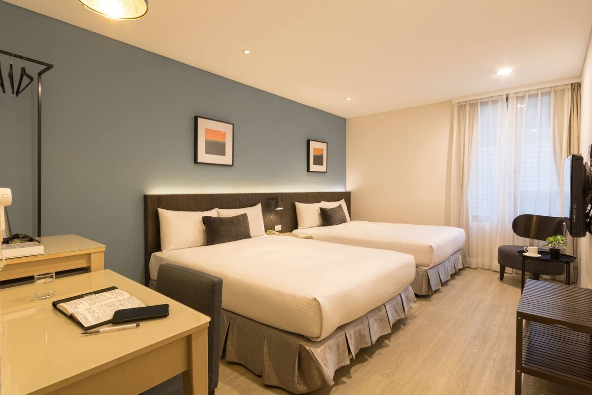 Habitación de hotel con dos camas, un escritorio, una silla, un televisor y una ventana con cortinas.