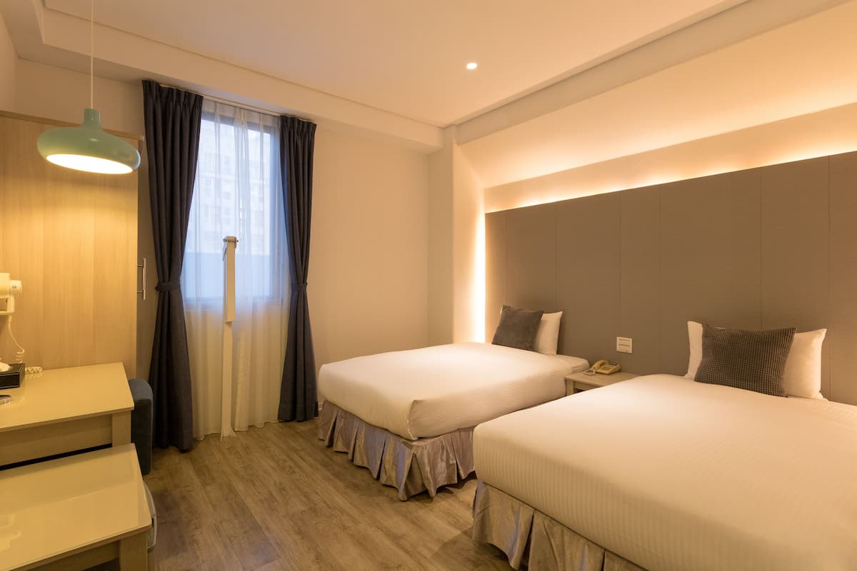 Habitación de hotel con dos camas, un escritorio, una lámpara y un ventanal con cortinas.