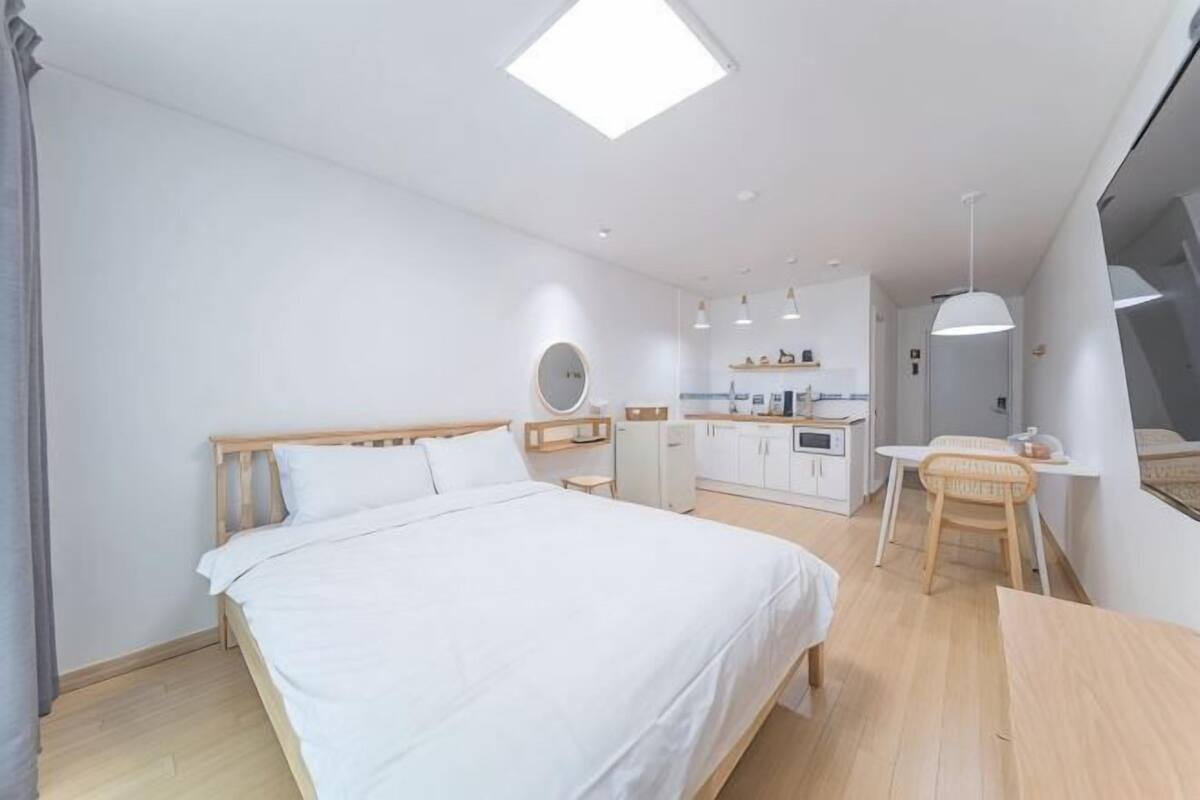 Un dormitorio moderno con una cama de madera, un escritorio pequeño, una silla y una kitchenette al fondo.