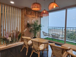 A modern interior with a dining area, wicker furniture, a large window with a view, and decorative elements like plants and wall art.
