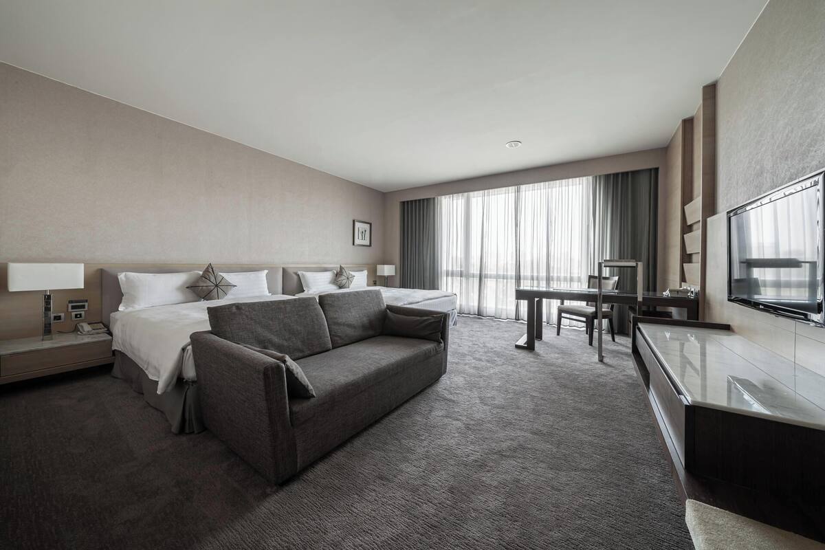 A hotel room with a bed, a sofa, a desk, a chair, a TV, and a large window with curtains.