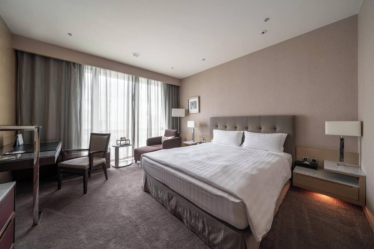 A hotel room with a large bed, a desk, a chair, a small table, and a window with curtains.
