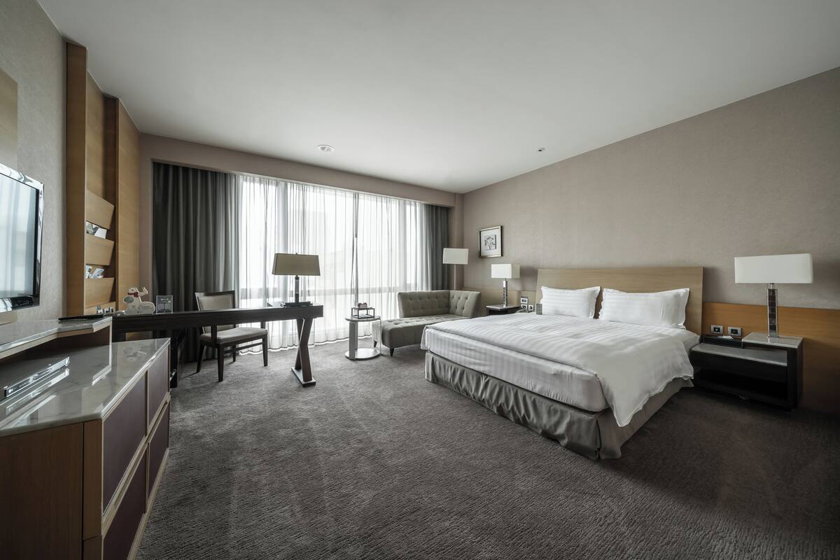 A hotel room with a large bed, a desk, a chair, a TV, and a desk lamp.