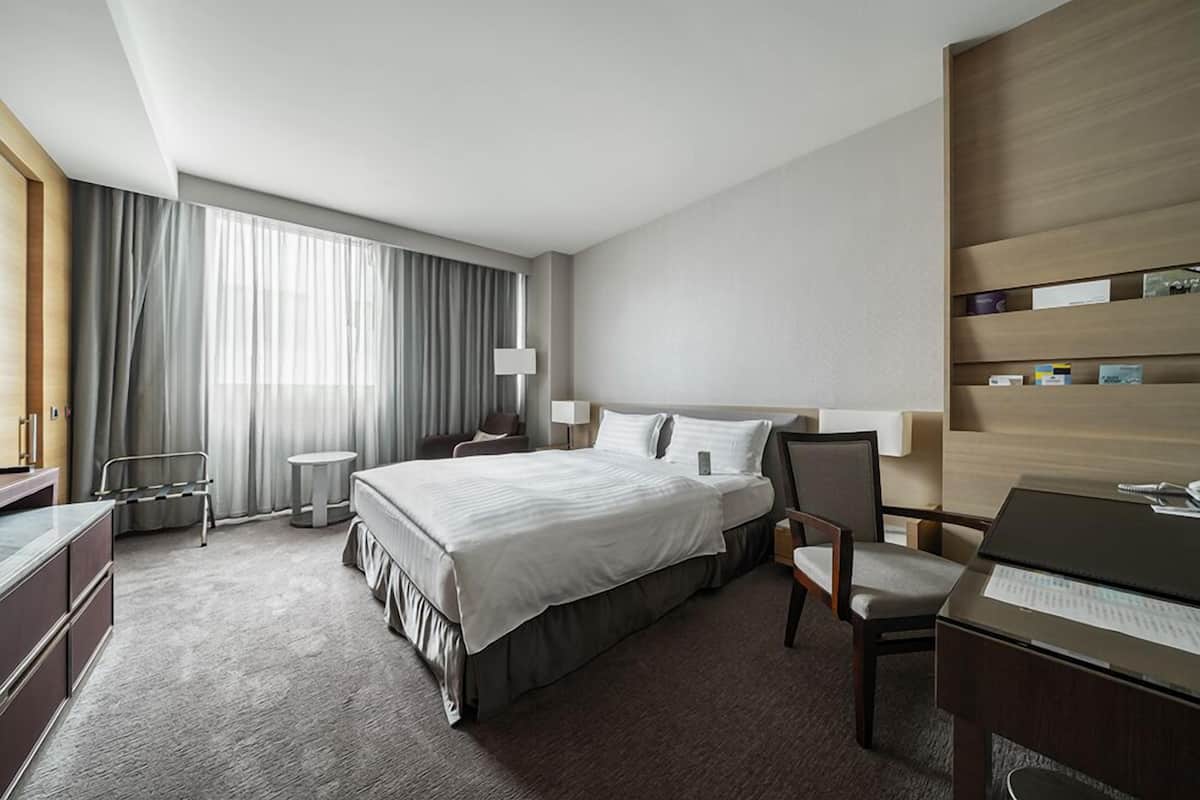 A modern hotel room with a large bed, a desk, a chair, and a bookshelf.