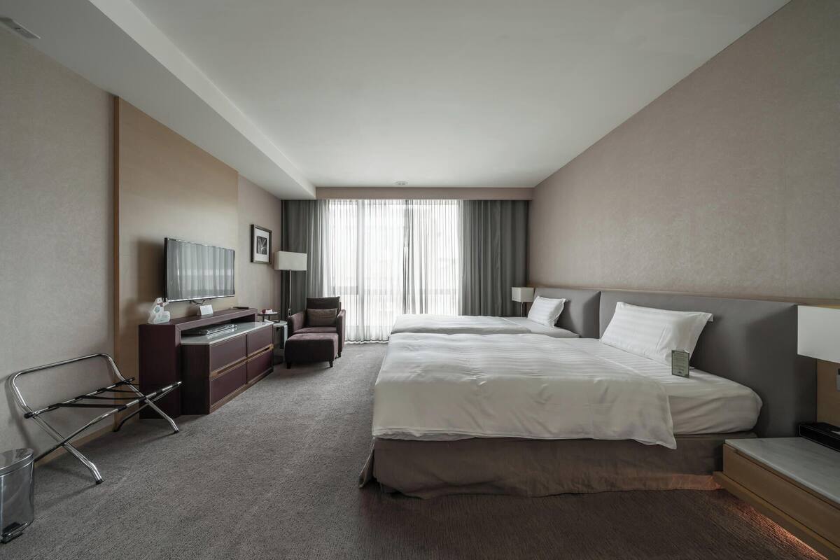 A hotel room with two beds, a desk, a chair, a TV, and a window with curtains.