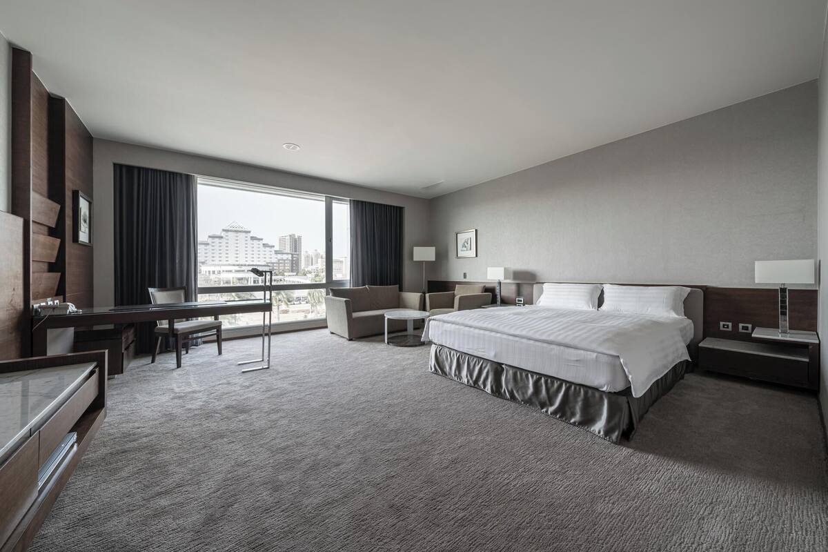 A hotel room with a large bed, a desk, a chair, a sofa, and a view of the cityscape through large windows.