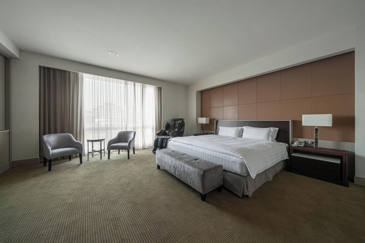 A hotel room with a large bed, two armchairs, a small table, and a nightstand with a lamp.