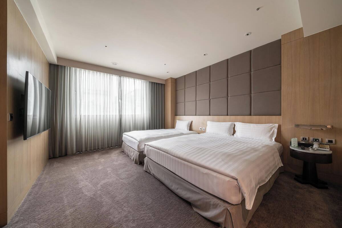 A modern hotel room with two beds, a flat-screen TV mounted on the wall, a bedside table with a lamp, and a large window with sheer curtains.