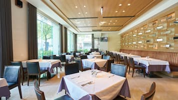 A spacious dining area with multiple tables and chairs, large windows, and a wooden ceiling.