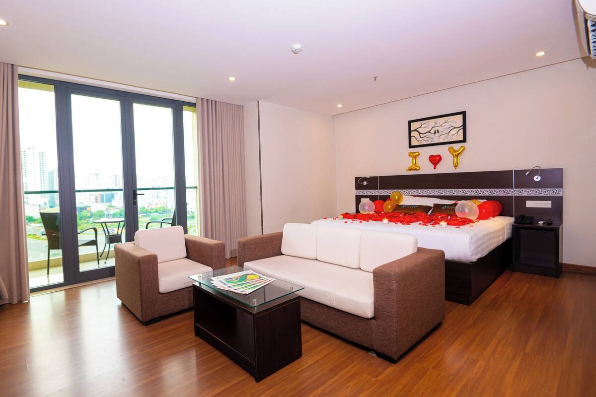 A modern hotel room with a large bed, a sofa, a coffee table, and a balcony with a city view.