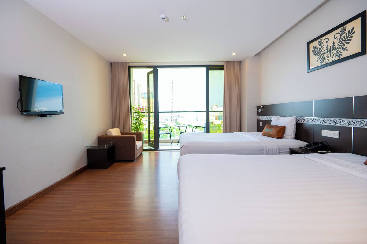 A hotel room with two beds, a TV, a desk, and a balcony with a view of the city.