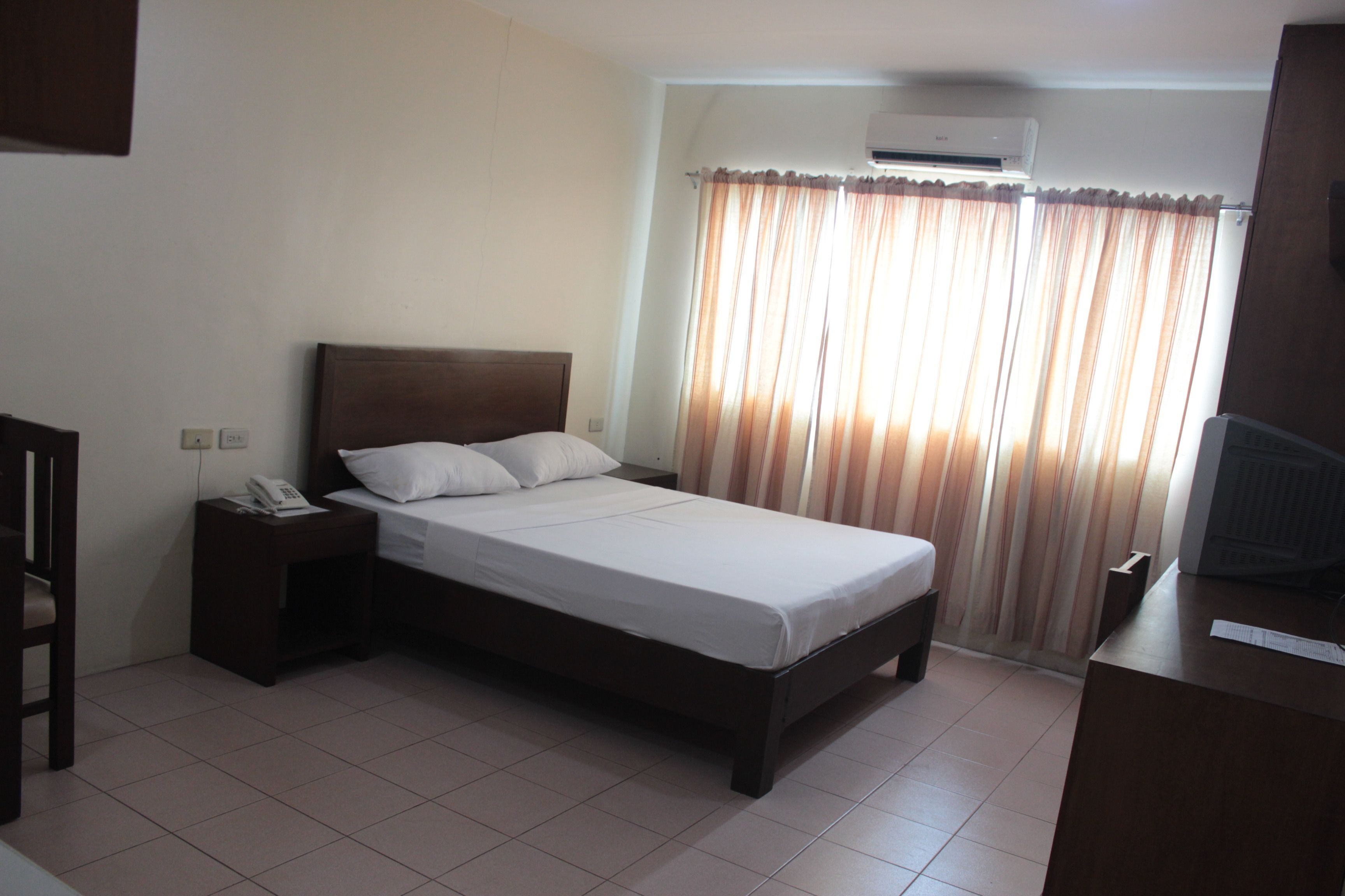 A hotel room with a bed, a nightstand with a telephone, a desk with a computer, and a window with curtains.