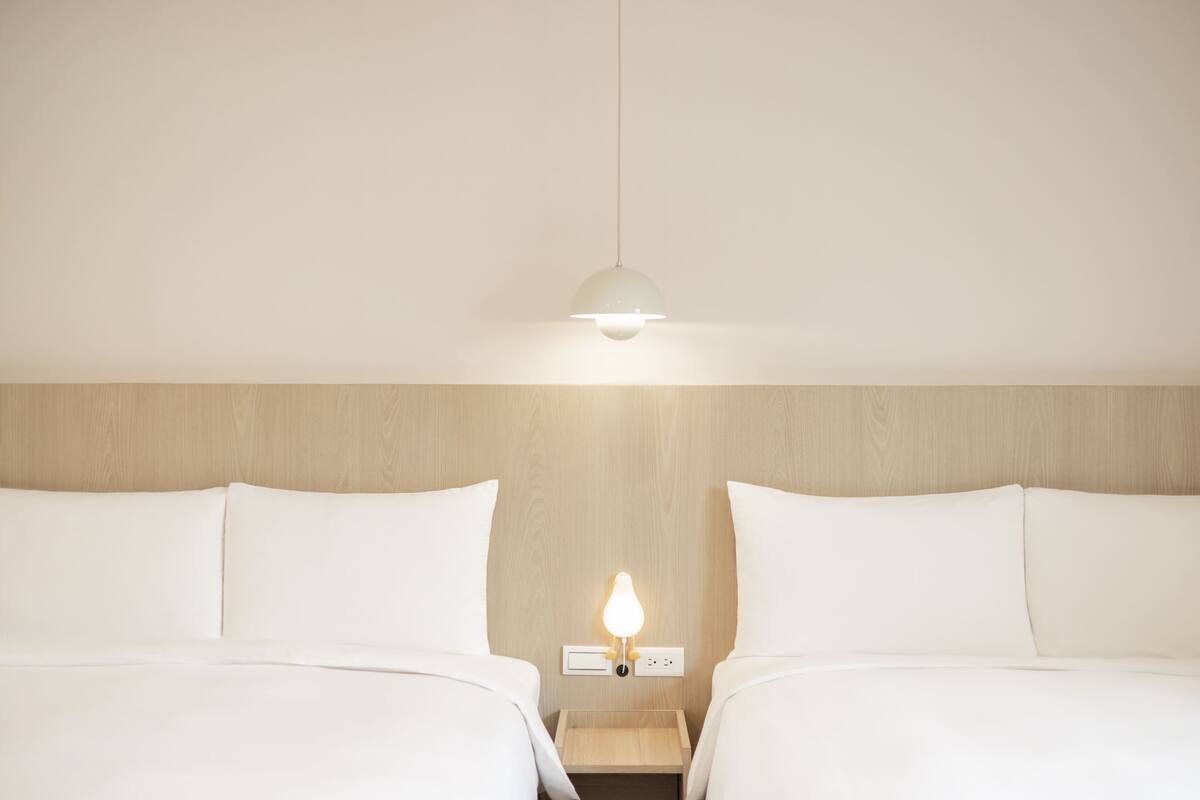 A hotel room with two beds, a wooden headboard, a bedside table with a lamp, and a hanging light fixture.