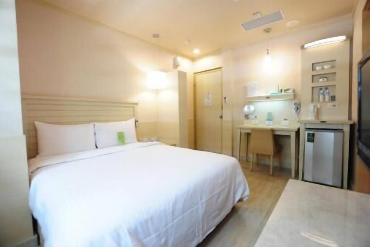 A hotel room with a bed, a desk, a chair, a small kitchenette, and a door.