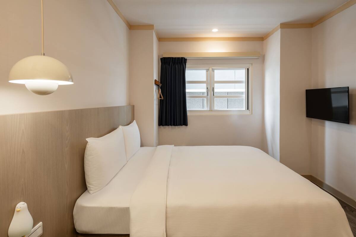 A hotel room with a large bed, a flat-screen TV mounted on the wall, a window with curtains, and a hanging light fixture.