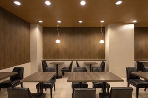 Un espace de restauration moderne avec des murs lambrés en bois, des spots encastrés au plafond et des chaises noires à revêtement en tissu disposées autour de tables carrées.