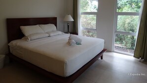 Una cama bien hecha con sábanas y almohadas blancas, una lámpara de noche y vistas a zonas verdes a través de amplias ventanas.