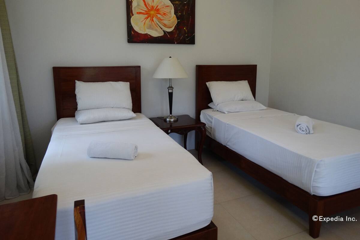 Una habitació d'hotel amb dos llits individuals, una meseta de nit amb una làmpada, un quadre amb un floreixement a la paret i una tovallola enrotllada sobre el llit.