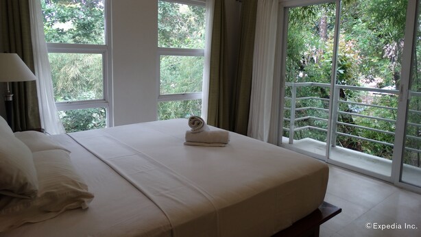 Habitación de hotel con una cama grande, una mesita de noche, una lámpara y vistas a zonas verdes por las ventanas.