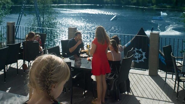 A deck with outdoor seating overlooking a river, with a wakeboarding ramp in the background. - Wake Valley Camping (Ylojarvi)