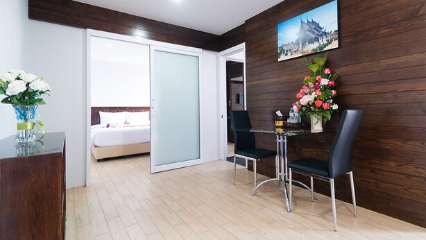 A hotel room with a bed, a desk with a vase of flowers, a dining table with chairs, and a wooden wall with a picture.