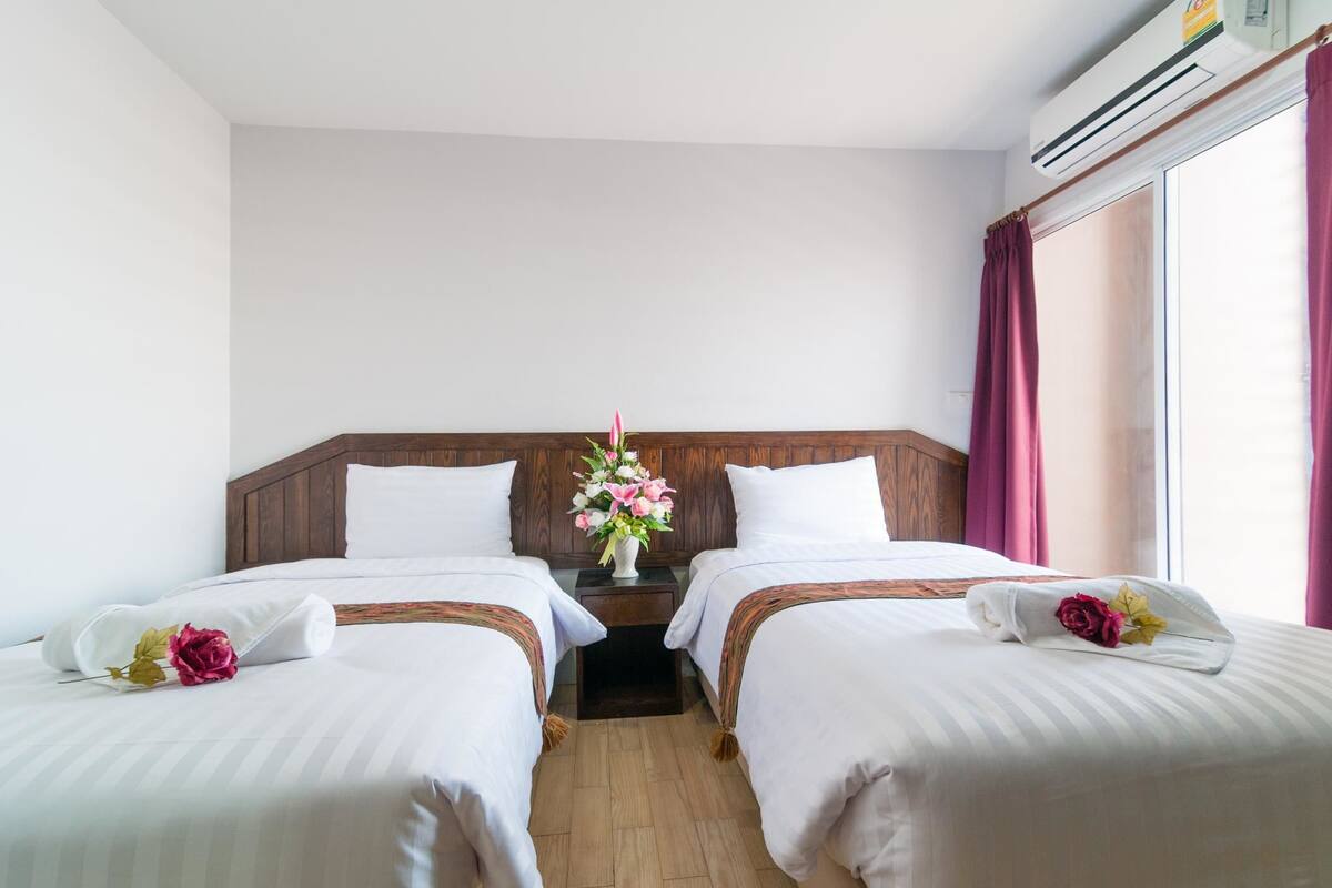 A hotel room with two beds, a wooden headboard, a nightstand with a vase of flowers, and a window with curtains.