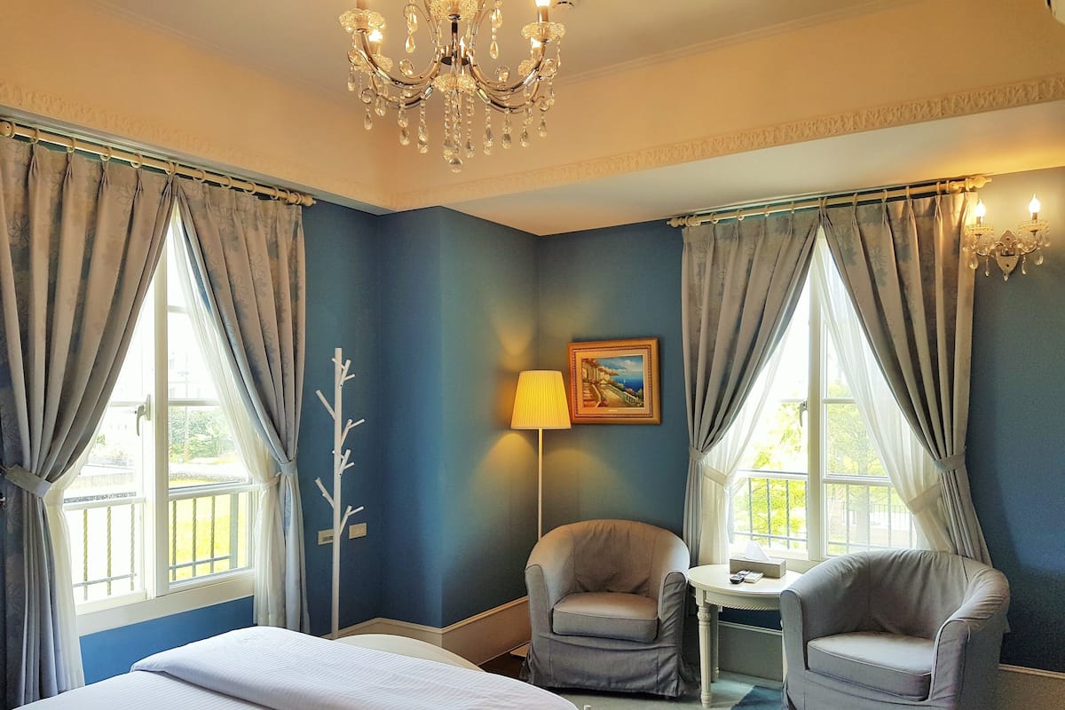 A hotel room with a large bed, two armchairs, a small table, a lamp, and a chandelier.