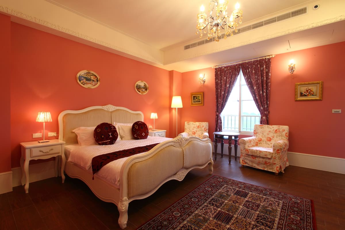 A bedroom with a large bed, two nightstands, a chair, a window with curtains, and a chandelier.