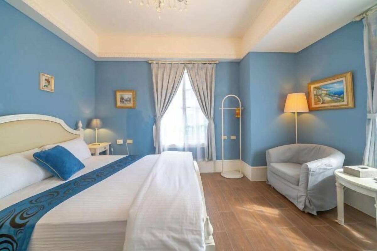 A hotel room with a bed, a chair, a bedside table, a lamp, and a window with curtains.
